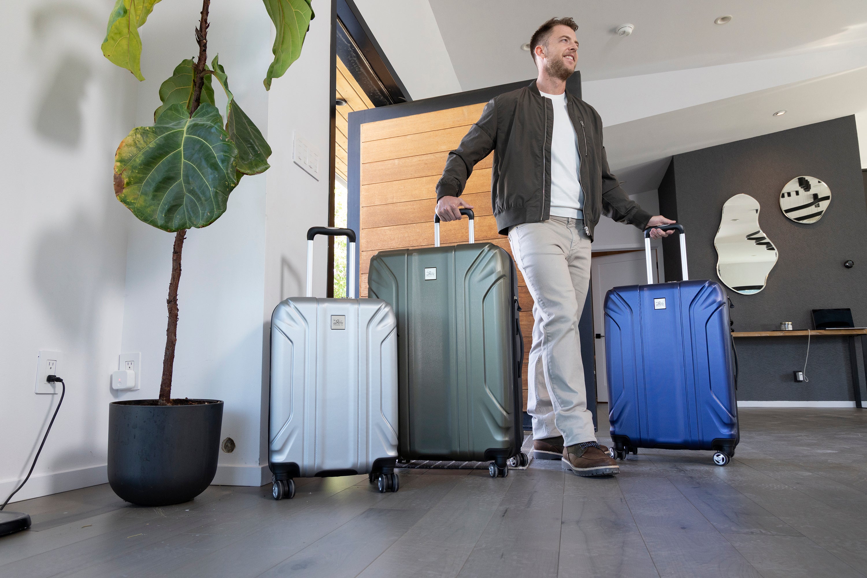 A man stands in a modern entryway with a three-piece Nimbus 5.0 luggage set in Silver, Olive Green, and Lazuli Blue lined up beside him, a wooden front door and fiddle leaf fig plant in the background.