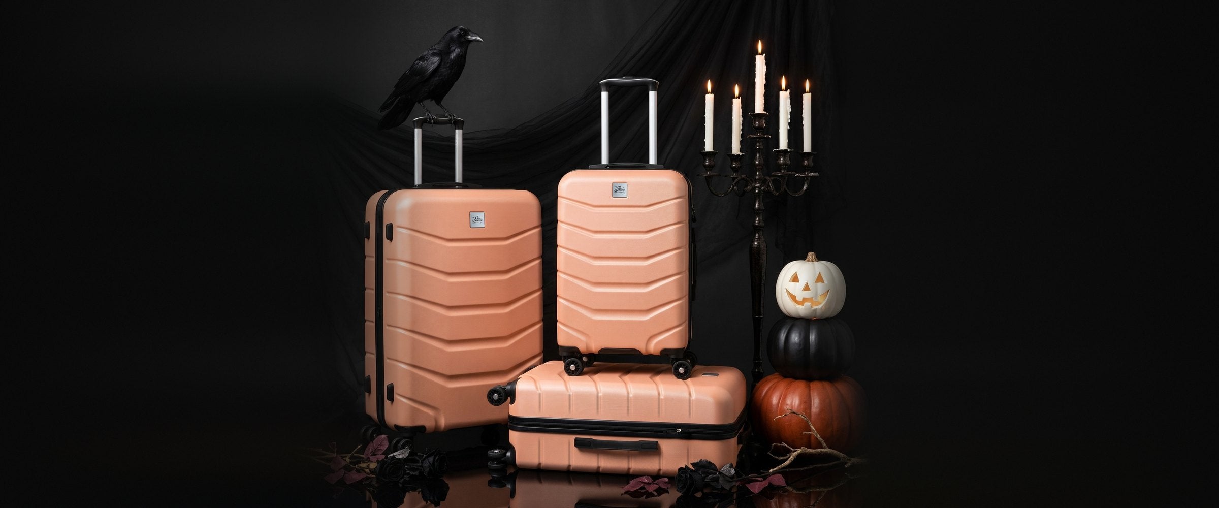Peach Skyway hardside luggage set with a raven perched on a handle, a lit candelabra and stacked Halloween pumpkins